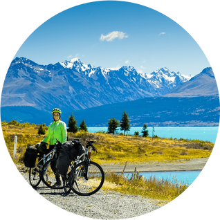 Cyclist stands on the winding gravel mountain road near Lake Pukaki view from Glentanner Park Centre near Mount Cook, on a background of blue sky with clouds, snowy Southern Alps